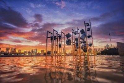 Cranes by river and buildings against sky at sunset