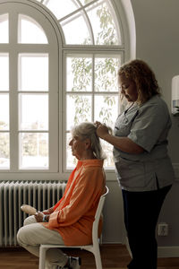 Side view of young female caregiver braiding senior woman's hair at nursing home