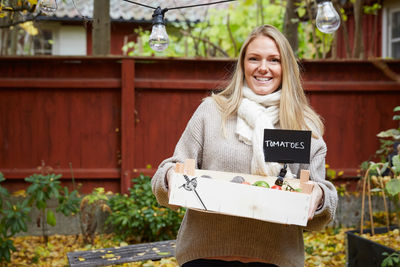 Portrait of smiling woman holding freshly produce tomato basket in yard