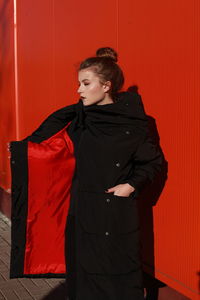 Fashionable teenage girl wearing black warm clothing standing against red wall during sunny day