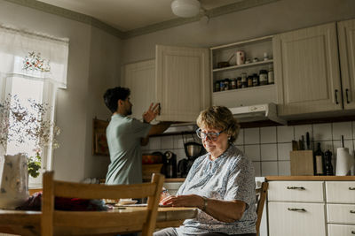 Elderly woman sitting at dining table while male caregiver working in kitchen