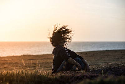 Woman sitting on shore at beach against sky during sunset