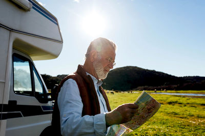 Side view mature male traveler examining paper map while standing near rv car during road trip in nature on summer day