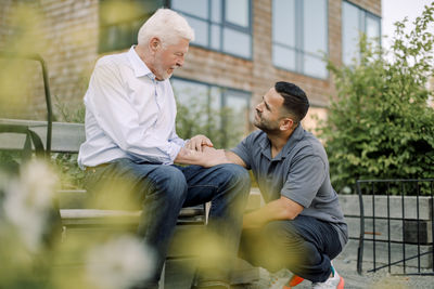 Male nurse squatting while talking to retired senior man sitting on bench at back yard