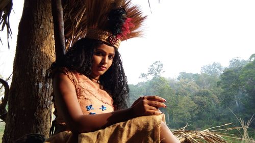 Beautiful young woman sitting on tree trunk against sky