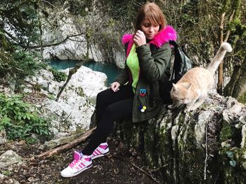 Woman looking at cat while sitting on rock against trees