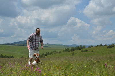 Man is playing with his dog on a meadow, surrounded with pink flowers and with hills in background