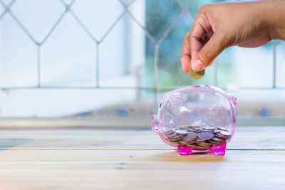 Cropped hand putting coins in piggy bank on table