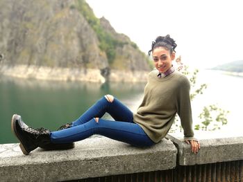 Portrait of smiling young woman sitting on retaining wall against lake