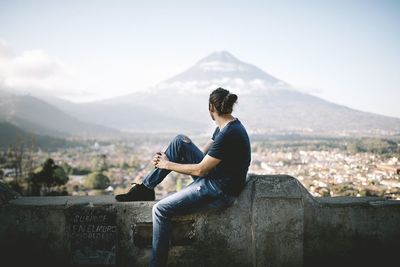 Side view of young man sitting on mountain against sky