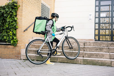 Full body female courier with thermal bag carrying bike on stairs towards building entrance while making delivery