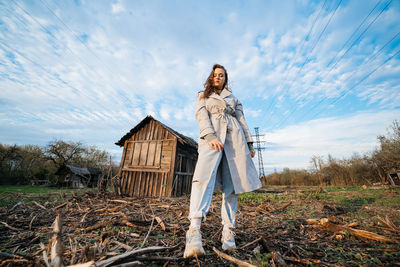 Beautiful girl with long hair in a grey trench coat next to an old wooden house blue sky background 