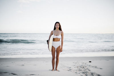 Full length portrait of young woman wearing bikini holding surfboard while standing at beach