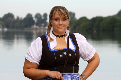 Portrait of smiling oktoberfest waitress standing with hand on hip against lake