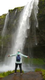 Rear view of woman standing with arms outstretched on rock by waterfall