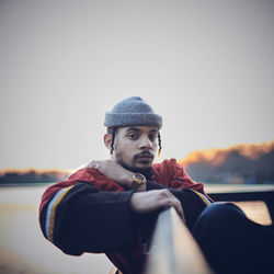 Portrait of young man sitting on bench against sky during sunset