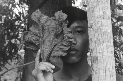 Portrait of man holding leaf by tree trunk in forest