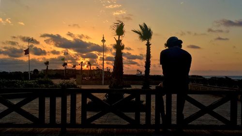 Silhouette man sitting on railing against cloudy sky during sunset