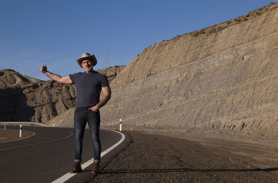 Adult man in cowboy hat hitchhiking on roadside on desert against mountain. almeria, spain