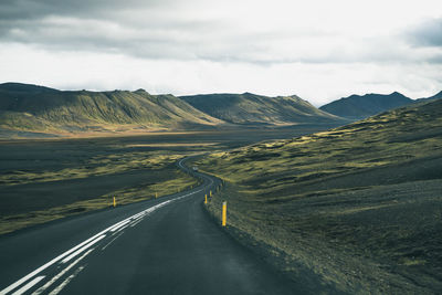 Country road leading towards mountains against cloudy sky