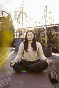 Smiling woman doing yoga while sitting cross-legged on exercise mat at terrace