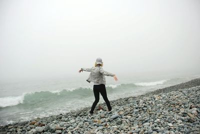Rear view of young woman with arms outstretched walking at beach against sky during foggy weather