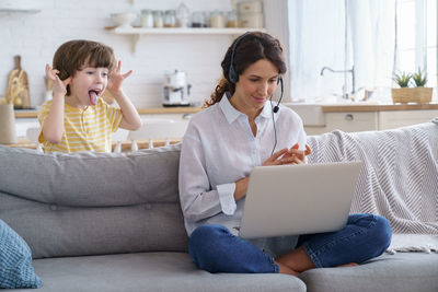 Mom talking on video call remote work on laptop from home with kid. children make noise shows tongue