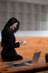 Asian business woman working at office and taking note, female sitting on desk and using laptop