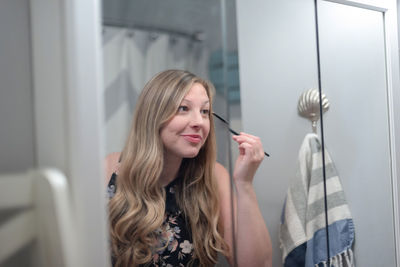 Reflection of smiling young woman applying make-up in mirror