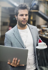 Mid adult man holding laptop and coffee in cafe