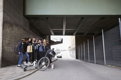 Teenage male and female friends with push scooters and bicycle on sidewalk below bridge