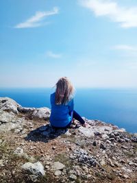 Rear view of woman sitting on rock by sea against sky