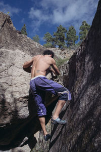 Rear view of shirtless man climbing on rock against sky
