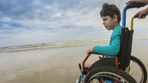 Side view of boy sitting in wheelchair at beach against sky