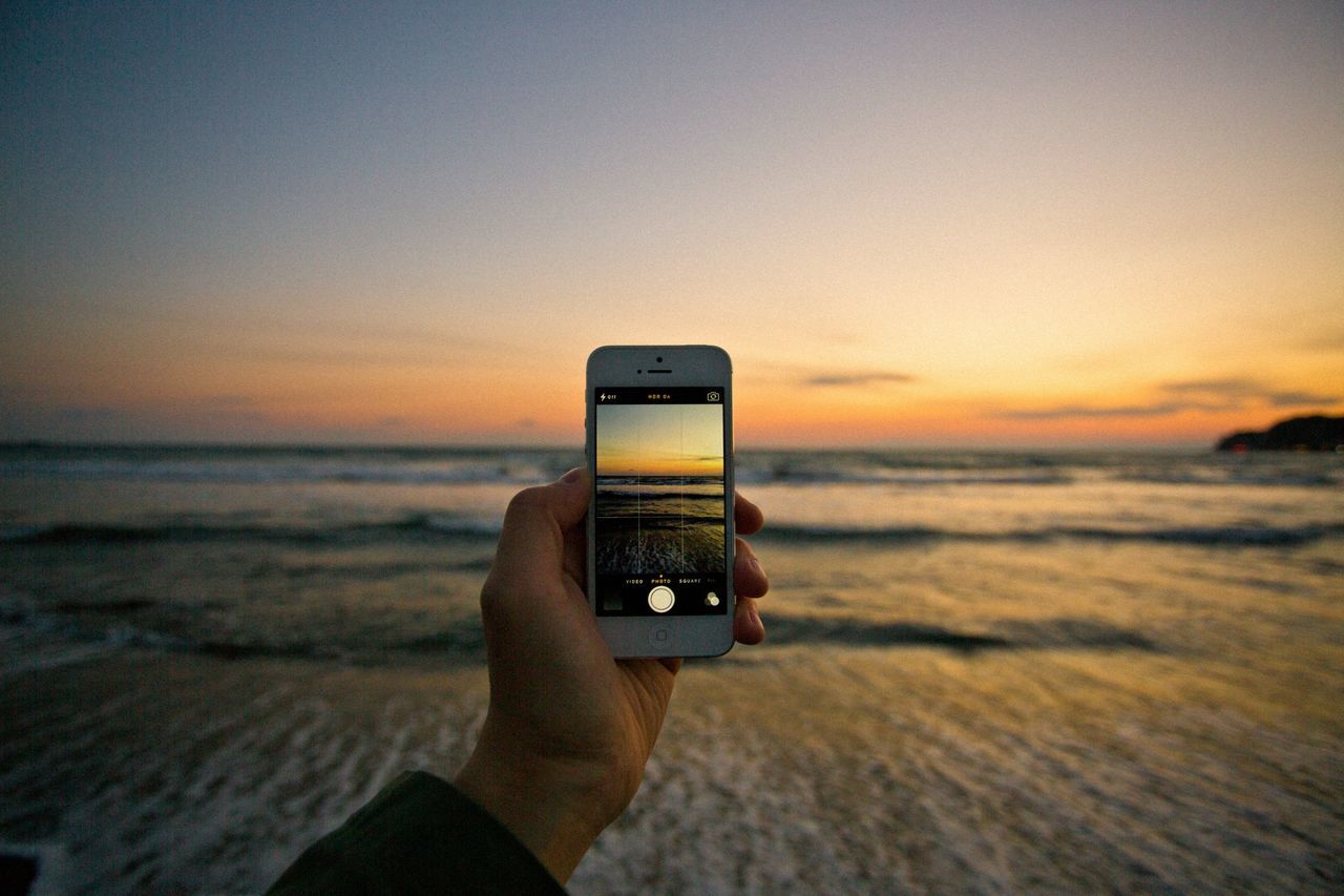 sunset, sea, horizon over water, holding, sky, person, part of, photography themes, technology, orange color, beach, cropped, focus on foreground, wireless technology, scenics, photographing, smart phone, beauty in nature