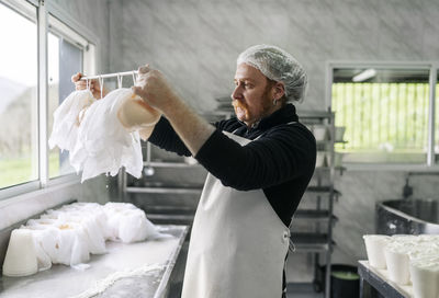 Mature male chef checking rod of hanging cheese wrapped in clothes at factory