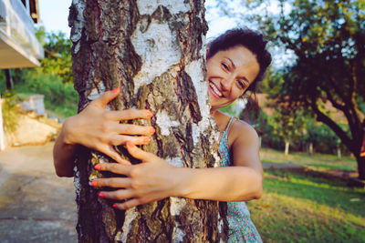 Portrait of smiling young woman embracing while standing by tree trunk