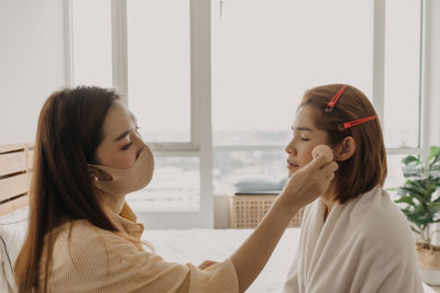 Side view of beautician wearing mask applying make up to woman