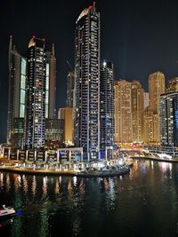 Illuminated modern buildings by river against sky at night