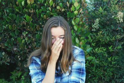 Shy teenage girl covering mouth with hand while standing against plants at park