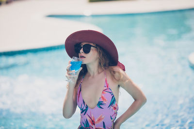 Woman in sunglasses and hat having drink against swimming pool