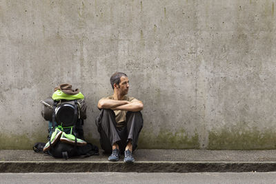Man sitting with backpack on footpath against wall