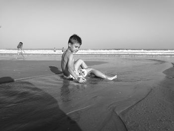 Full length of shirtless boy sitting on shore at beach against sky