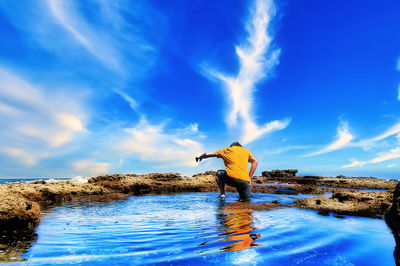 Man standing on rock by sea against blue sky