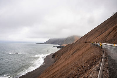 View of steep brown rocky slope beside the ring road by beach against cloudy sky