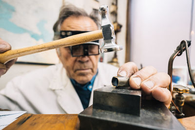 Focused skilled mature male master in glasses using hammer and chisel while working with gemstone in jewelry workshop