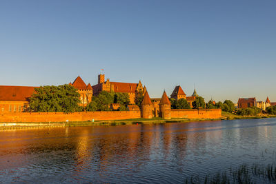 Buildings by lake against a clear blue sky, malbork castle. 