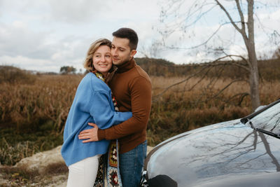 A man and a woman embrace happily in front of a car, smiling under the sky