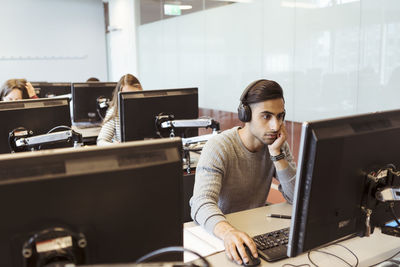 Young male student wearing headphones while using computer at university library