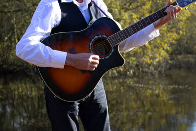 Midsection of man playing guitar while standing against lake